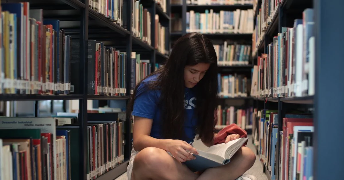 Une jeune femme assise par terre dans une bibliothèque, lisant un livre avec attention.