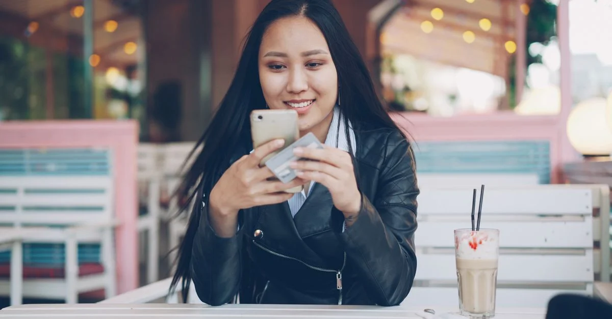 Femme souriante dans un café utilisant son smartphone et sa carte de crédit pour le shopping en ligne.
