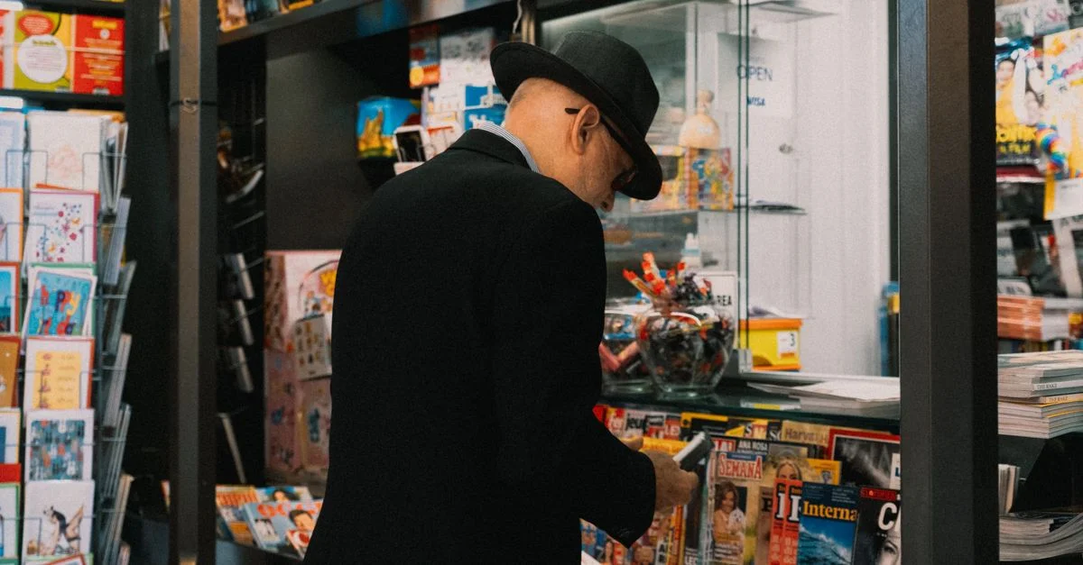 Un homme senior portant un chapeau feuilletant des magazines à un kiosque. Cadre intérieur avec des médias imprimés divers.