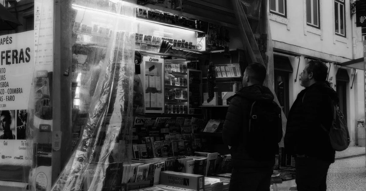 Photo en noir et blanc de deux hommes près d'un kiosque de rue, créant une atmosphère urbaine classique.
