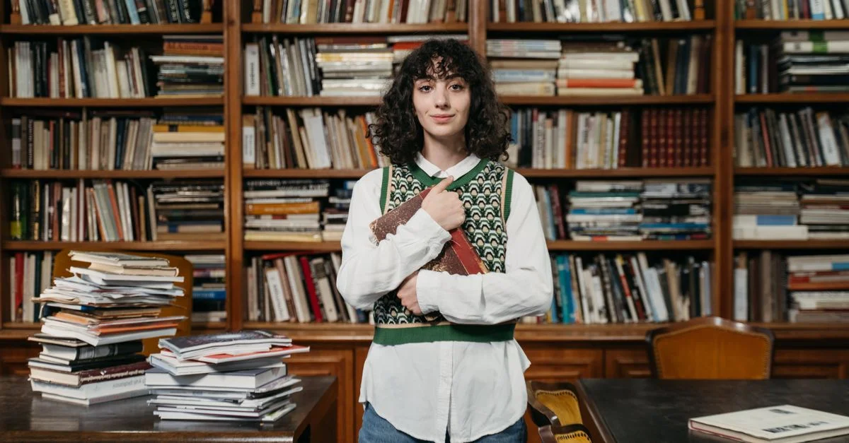 Une femme se tient avec assurance dans une bibliothèque, entourée d'étagères, tenant un livre.